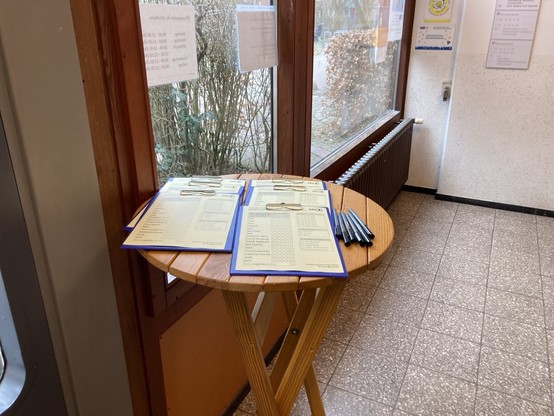 Ein runder Holstisch mit blauen Klemmbrettern und Filzstiften daneben. Auf den Klemmbrettern klemmt jeweils ein Zettel. Dort kann man ankreuzen, für welche Partei man heute die 5 Stimmmen der Landesliste verteilt hat und zusätzlich statistische Angaben über sich selbst.